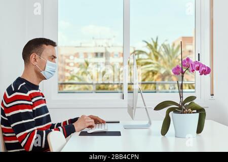 closeup of a young caucasian man indoors, wearing a surgical mask, using a computer sitting at a white table Stock Photo