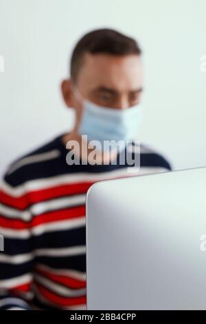 a young caucasian man indoors, wearing a surgical mask, using a computer, with the focus in the computer in the foreground Stock Photo