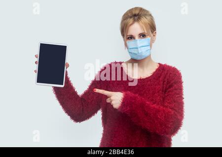 Portrait of beautiful young woman with surgical medical mask in red blouse standing and holding tablet empty screen and pointing finger to device and Stock Photo