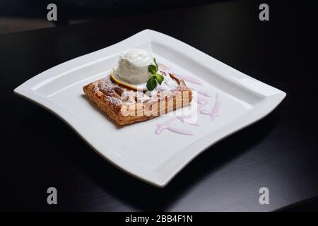 Delicious dessert served with fresh mint and powdered sugar on white plate in restaurant. From above view of piece of crusty cake with white vanilla ice cream on fruit slice on top. Stock Photo