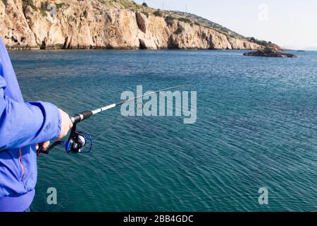 a fisherman's hand in a blue jacket holds a spinning rod over the greenish sea water next to rocks and fishes Stock Photo