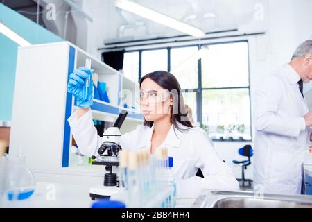 Laboratory, biotechnology, experiment. Young brunette cute intern is studying new technology in science, she is wearing labcoat and safety glasses Stock Photo