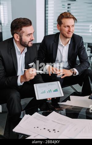 two young partner business man investor talking and reading information about financial statistics in mobile tablet together sitting in office, financ Stock Photo