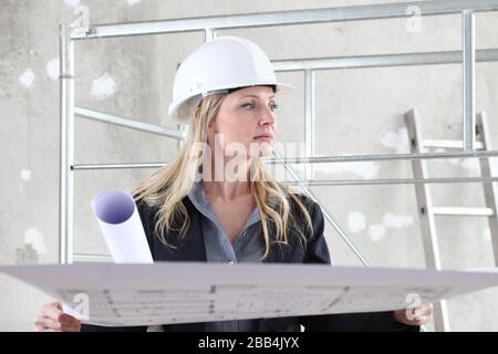 woman architect or construction engineer reading blueprint wear helmet inside a building site with scaffolding in the background Stock Photo