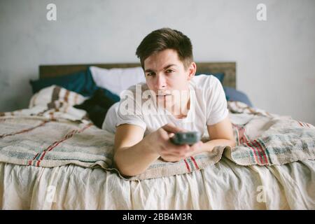 Guy using remote control while sitting on the bed in home. Happy attractive young man watching tv . Human emotions on her face. Stock Photo