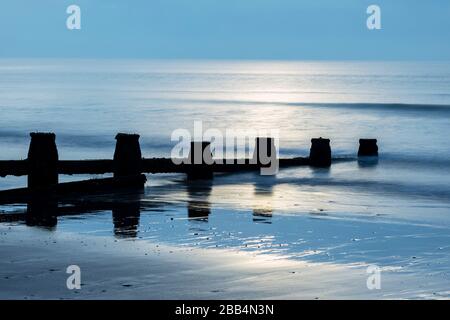 Sun rising over the ocean with water rushing around timbe groynes on Dawlish Warren beach Stock Photo