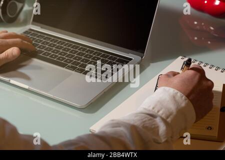 The hands of a businessman man who uses the laptop and takes notes working from home. Note pad and pen on the table. Telework concept Stock Photo