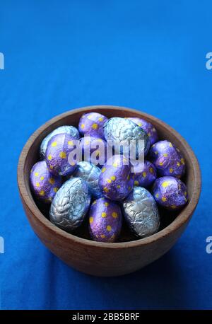 A wooden bowl filled with colorful chocolate Easter eggs in foil wrappings, with a blue table cloth background. High angle view and copy space above. Stock Photo