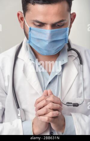 Close up portrait of mature Indian male medical doctor in uniform praying, standing on plain background.concept of cold and flu, fever and coronavirus Stock Photo