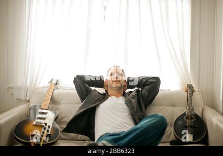 Mid-adult man sitting on a couch with two guitars in his living room. Stock Photo