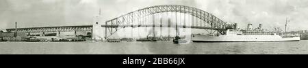 A vintage 1930s panoramic view of Sydney Harbour Bridge looking west in November 1931 when the bridge was nearing completion. The P&O ocean liner 'Strathnaver' on her maiden voyage from London is being guided by tug boats below it. Sydney Harbour Bridge is a steel arch bridge. It was designed and built by Dorman Long of Middlesbrough, England. The last stone of the north-west pylon (shown here on right) was placed in January 1932. It was opened on 19 March 1932. P&O's RMS Strathnaver was part of the Royal Mail Ships that worked the route between the UK and Brisbane, Queensland, Australia. Stock Photo