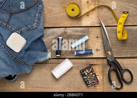 Torn blue denim cotton fabric trousers with tailor's tools on brown wood background - jeans fashion mending or repair concept, flat lay top view from Stock Photo