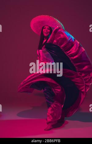 Fabulous Cinco de Mayo female dancer on purple studio background in neon light. Beautiful female model in traditional costume and sombrero dancing. Celebration, holiday, beauty and fashion concept. Stock Photo