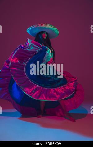 Fabulous Cinco de Mayo female dancer on purple studio background in neon light. Beautiful female model in traditional costume and sombrero dancing. Celebration, holiday, beauty and fashion concept. Stock Photo