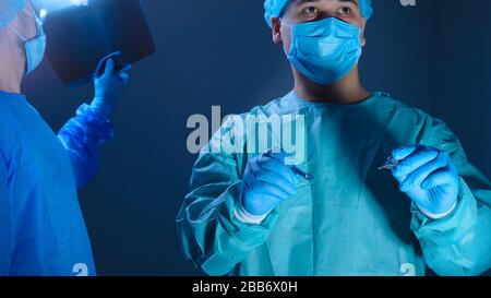 Portrait of two surgeons in sterile gowns, hats and masks. One surgeon, preparing to operate, holds a scalpel with a needle holder in his hands, anoth Stock Photo