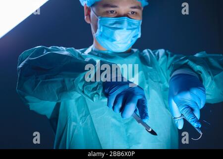 close-up portrait of a caucasian doctor surgeon, in a sterile suit, mask, holding a scalpel and needle holder with a needle, guiding them to the opera Stock Photo