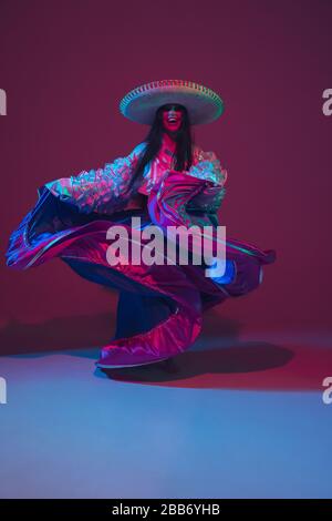 Fabulous Cinco de Mayo female dancer on purple studio background in neon light. Beautiful female model in traditional costume and sombrero dancing. Celebration, holiday, beauty and fashion concept. Stock Photo