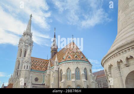 The spire of the famous Matthias Church in Budapest, Hungary. Roman Catholic church built in the Gothic style. Orange colored tile roof. Blue sky and white clouds above. Horizontal photo with filter. Stock Photo
