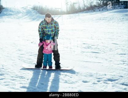 Father teaches daughter how to ride on snowboard. Have fun together standing on snowboard. Winter holidays in mountain during sunny day. Ground full o Stock Photo