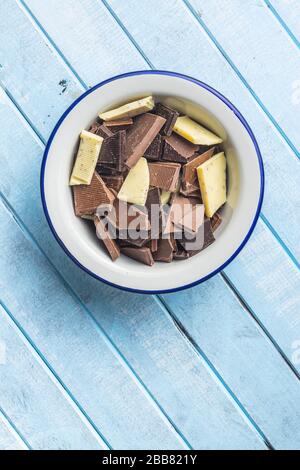 Broken white, milk and dark chocolate bars in bowl on blue wooden table. Stock Photo