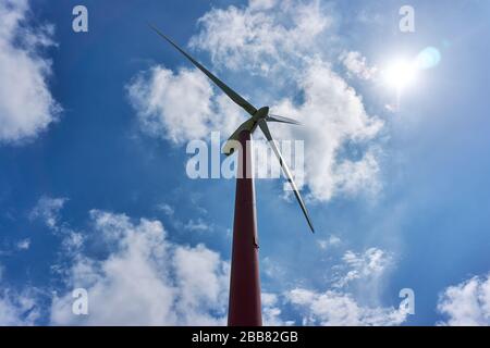 Wind turbine against saturated dramatic blue sky with white clouds, sun and lens flare. Low angle view of red and white painted wind turbine. Stock Photo