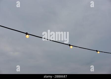 String light many Yellow glowing outdoor festival string light bulbs against a dark grey stormy and cloudy sky with copy space. Stock Photo