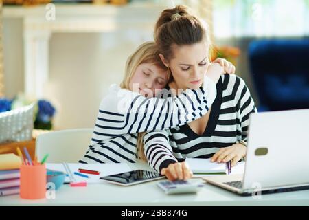 child craving attention while mother busy working in home office in the modern house in sunny day. Stock Photo