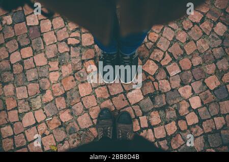 View from above. Two pairs of people legs standing on road of tiles or orange pavement. Wear black pants and shoes or sneakers Stock Photo