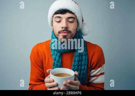 Young bearded man isolated over background. Calm peaceful guy with white cup in hands and closed eyes. Scarf around neck. Wear red restive winter clot Stock Photo