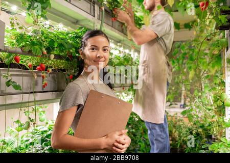 Portrait of smiling confident young female worker of small greenhouse standing at plant shelf and holding clipboard Stock Photo