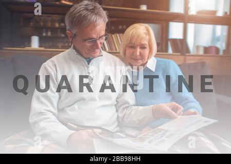 Beautiful old couple are sitting on the couch together and reading a big newspaper. Man is using reading glasses. They look concentrated and consious Stock Photo