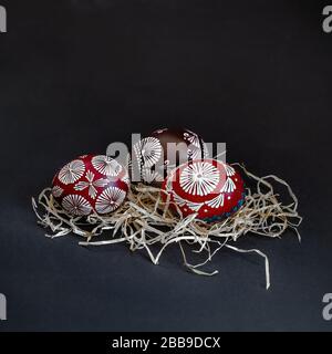 Three traditionally Lithuanian painted red, black, white, blue eggs with wax, called marguciai, in the straw nest on a black background. Copy paste Stock Photo