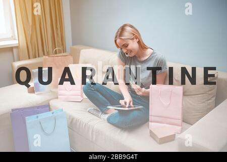 Online shopping at home. Young smiling shopper with tablet and credit cards is ordering by smarthphone in online shop while sitting on a brown sofa wi Stock Photo