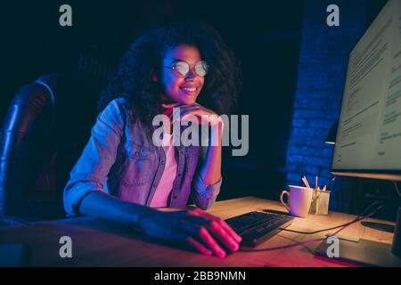 Profile photo of pretty cheerful dark skin programmer lady look monitor check debugging system work overtime read email wear specs casual shirt sit Stock Photo