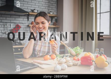 Cheerful nice attractive woman sit at table in kitchen. She smile and put wire headphones in ears. Model look on laptop. Vegetables lying on table Stock Photo
