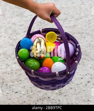 A purple easter egg basket with shiny colored plastic easter eggs in it being held by a child's hand. Stock Photo