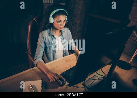 Photo of young dark skin business lady look big screen work overtime use modern earphones listen voice messages wear specs casual denim shirt sitting Stock Photo