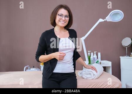 Beautiful smiling mature woman client of beauty salon, posing looking at camera, background massage table and equipment. Beauty industry, skin care, h Stock Photo