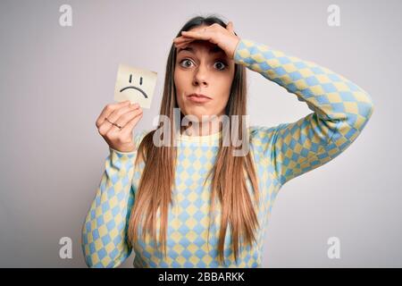 Young blonde woman holding sticky paper note with sad face emoticon over isolated background stressed with hand on head, shocked with shame and surpri Stock Photo