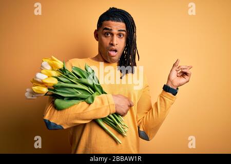 Young african american afro romantic man with dreadlocks holding bouquet of yellow tulips Surprised pointing with finger to the side, open mouth amaze Stock Photo