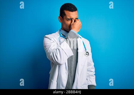Handsome african american doctor man wearing coat and stethoscope over blue background tired rubbing nose and eyes feeling fatigue and headache. Stres Stock Photo