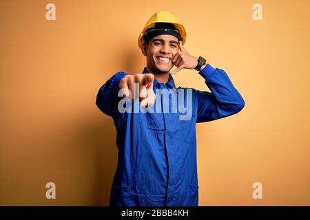 Young handsome african american worker man wearing blue uniform and security helmet smiling doing talking on the telephone gesture and pointing to you Stock Photo