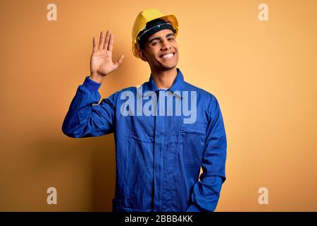 Young handsome african american worker man wearing blue uniform and security helmet Waiving saying hello happy and smiling, friendly welcome gesture Stock Photo