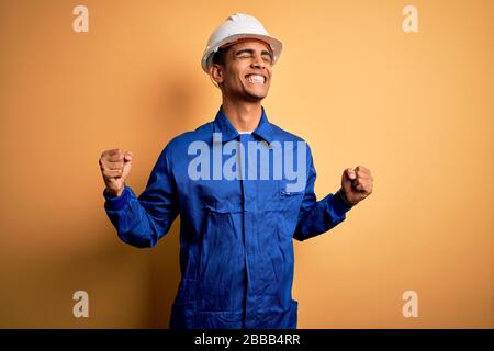Young handsome african american worker man wearing blue uniform and security helmet very happy and excited doing winner gesture with arms raised, smil Stock Photo