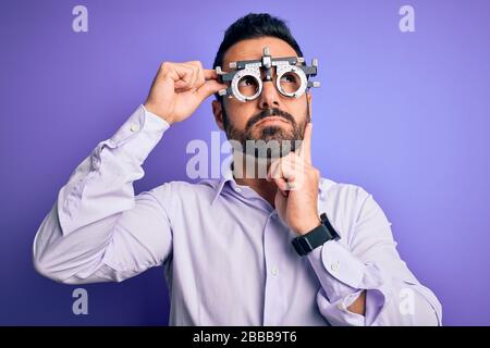 Young handsome optical man with beard wearing optometry glasses over purple background serious face thinking about question, very confused idea Stock Photo