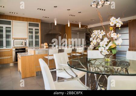Round glass dining table with white leather and chrome armchairs, wood and glass cabinets and island with grey nuanced granite countertop and white leather high back chairs in kitchen with grey marble tile floor inside a modern luxurious multistory penthouse condominium unit, Old Montreal, Quebec, Canada. This image is property released for publication in calendars and editorial use. EUPR0359