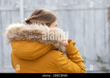 Back view of woman wearing yelllow furry parka jacket in front of grey wooden background Stock Photo