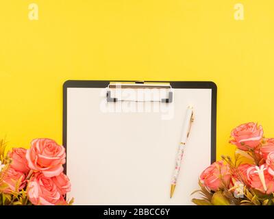 Clipboard with blank copy space, pen on it, pink roses flowers around on yellow background. Flat lay, top view mock up. Stock Photo