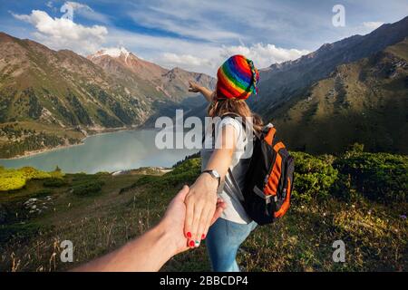 Tourist woman in rainbow hat and brown poncho holding man by hand and pointing to the lake in the mountains Stock Photo