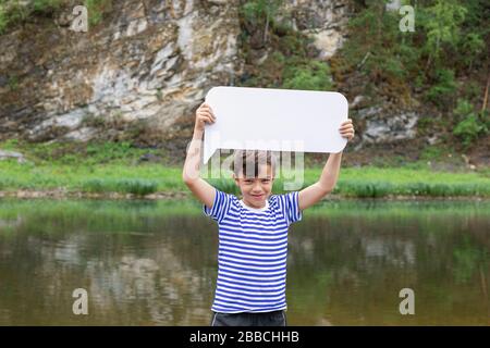 Little smiling boy in striped t-shirt standing on bank of river keeping blank white board in hands above his head, rocky cliffs on background. Summer Stock Photo